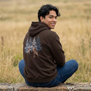 May include: A dark brown hooded sweatshirt featuring a graphic on the back. The design displays a detailed illustration of a deer standing in front of a group of blue-toned pine trees. The hoodie has long sleeves and a hood with a drawstring.