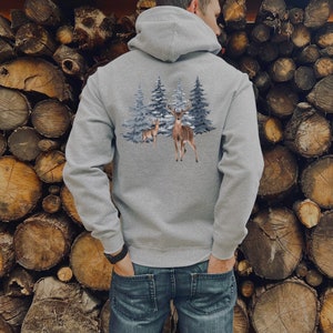 May include: A heather gray hooded sweatshirt featuring a graphic print on the back. The design displays two brown deer standing in front of a cluster of blue-toned evergreen trees. The hoodie has long sleeves, a hood, and ribbed cuffs.