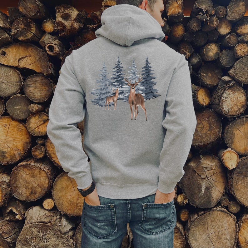 May include: A heather gray hooded sweatshirt featuring a graphic print on the back. The design displays two brown deer standing in front of a cluster of blue-toned evergreen trees. The hoodie has long sleeves, a hood, and ribbed cuffs.