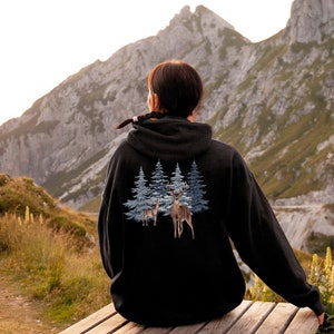 May include: A black hooded sweatshirt featuring a graphic print on the back. The design displays two brown deer standing in front of a cluster of blue-toned evergreen trees. The hoodie has long sleeves and a hood, shown from a rear perspective.