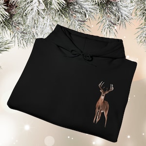 May include: A black hooded sweatshirt featuring a graphic of a brown deer with antlers positioned on the lower right side. The garment includes a hood with matching black drawstrings and long sleeves, presented in a folded, flat-lay orientation.