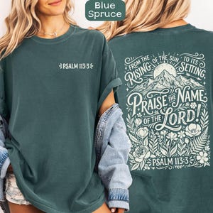 May include: Blue Spruce t-shirt with white text. The front of the shirt has "PSALM 113:3". The back of the shirt has the text "FROM THE RISING OF THE SUN TO ITS SETTING, PRAISE THE NAME OF THE LORD! PSALM 113:3" with a mountain and floral design.