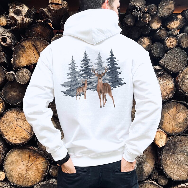 May include: A white hooded sweatshirt featuring a graphic print on the back. The design displays a large buck with antlers and a smaller deer standing in front of a cluster of dark grey evergreen pine trees against a plain white background.