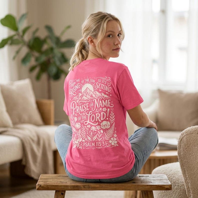 May include: A pink t-shirt with white text on the back. The text reads "From the rising of the sun to its setting. Praise the name of the Lord! Psalm 113:3" with a mountain and floral design. The shirt is worn by a person.