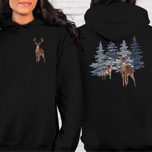 May include: A black hooded sweatshirt featuring a small graphic of a brown deer on the front left chest. The back of the hoodie displays a larger graphic of two brown deer standing in front of a cluster of blue-toned evergreen pine trees.
