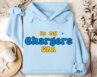 May include: A light blue hooded sweatshirt featuring the text "In my Chargers ERA" printed in a bold, retro-style font. The words "In my" and "ERA" are in yellow, while "Chargers" is in blue, accented by small yellow and blue star icons.