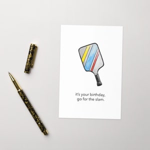 May include: A white greeting card with a watercolor illustration of a pickleball paddle with yellow, blue, and red stripes. The text reads, "it's your birthday, go for the slam." A black and gold pen is next to the card.