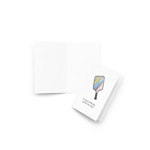 May include: A white greeting card with a colorful pickleball paddle illustration. The paddle has yellow, blue, and red stripes. Text on the card reads, "It's your birthday, go for the slam."