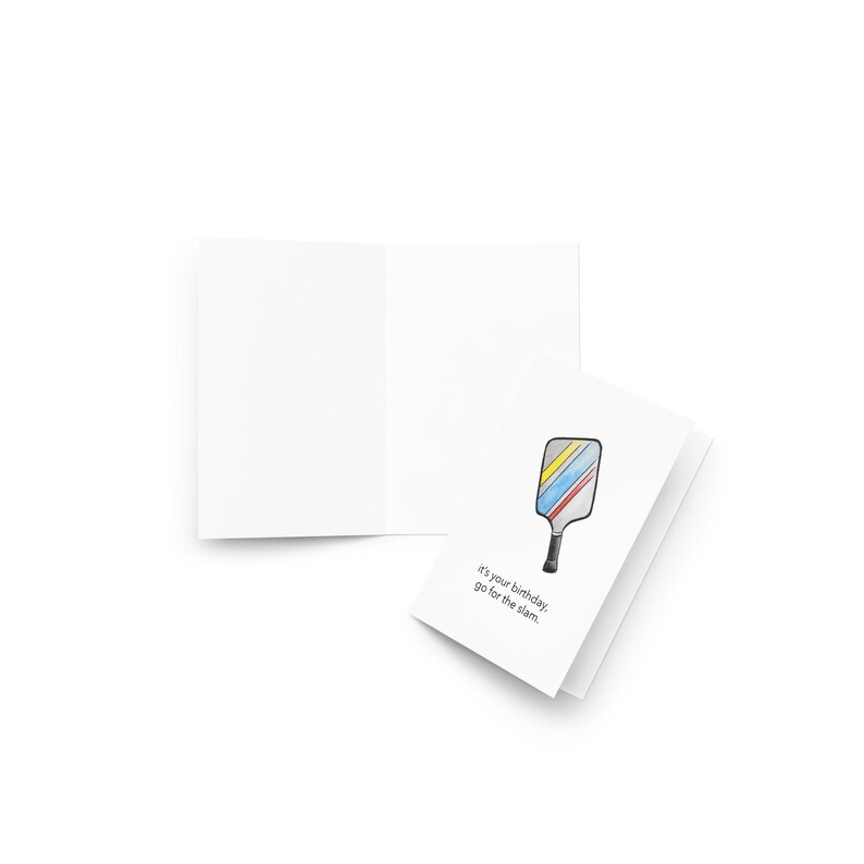 May include: A white greeting card with a colorful pickleball paddle illustration. The paddle has yellow, blue, and red stripes. Text on the card reads, "It's your birthday, go for the slam."