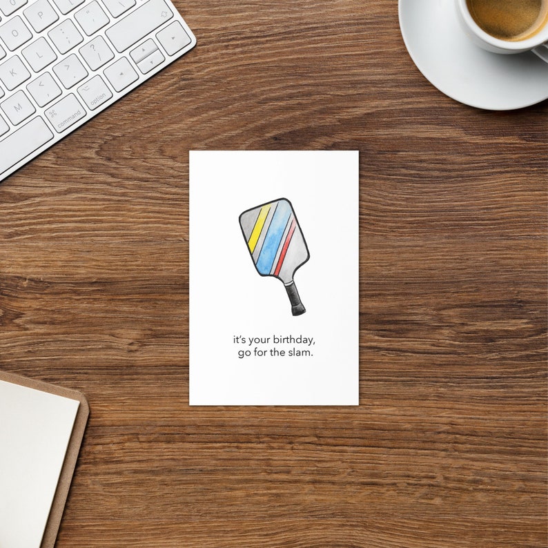 May include: A birthday greeting card featuring a watercolor illustration of a pickleball paddle with blue, yellow, and red stripes. The card includes the text "it's your birthday, go for the slam."