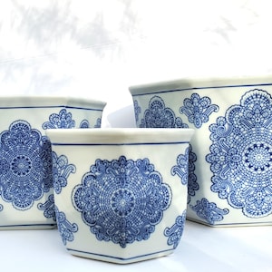 May include: Three white ceramic planters with a blue floral design. The planters are hexagonal and vary in size, with the largest being approximately 12 inches tall. The blue pattern is intricate and covers the sides of the planters.