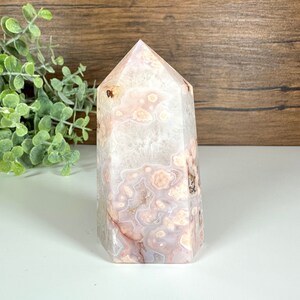 May include: A polished crystal tower with a flat base and a pointed top. The crystal features a mix of white, pink, and brown patterns. The crystal is standing on a white surface with a wooden background.