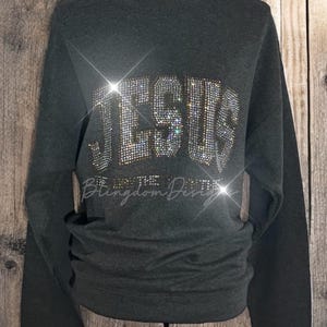 May include: A dark green sweatshirt with the word "JESUS" spelled out in rhinestones. The sweatshirt has long sleeves and a crew neck.
