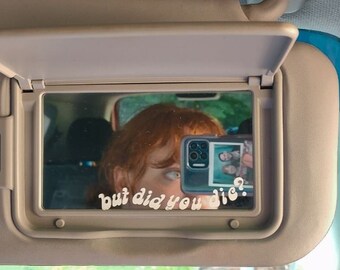 May include: A car visor with a mirror. The mirror reflects a person with red hair and a phone displaying a photo. The mirror has the text "but did you die?" in a handwritten font. The visor is beige.