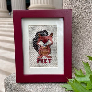 May include: A framed cross-stitch featuring a cartoon beaver with the word "MIT" underneath. The frame is a dark red color.