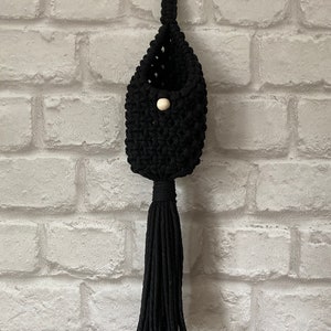 May include: Black macrame hanging planter with a wooden ring at the top and a wooden bead at the bottom. The planter has a tassel.