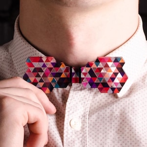 Könnte beinhalten: Eine geometrische Fliege mit einem farbenfrohen Dreiecksmuster in Rot, Lila, Orange und Blau. Die Fliege wird auf einem hellrosa Button-Down-Hemd getragen. Die Hand der Person justiert die Fliege.