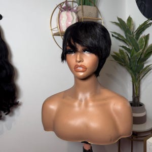 Peut inclure: Une perruque courte noire de style pixie avec une frange sur une tête de mannequin. La perruque est coiffée avec une raie sur le côté et la frange est coupée droite sur le front.