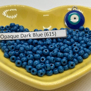 Peut inclure: Un plat en forme de cœur jaune rempli de perles de verre bleu foncé opaque. Les perles sont petites et rondes. Un charme argenté avec un motif d'œil bleu maléfique se trouve sur le côté droit du plat. L'étiquette sur le plat indique "Opaque Dark Blue (615)".
