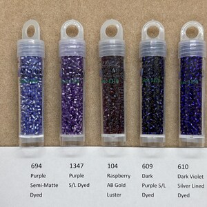 May include: Six small glass bottles filled with seed beads in various colours, including purple, dark purple, raspberry, dark blue, and dark violet. The bottles are labelled with numbers and descriptions of the bead colours and finishes.