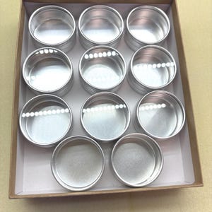 Peut inclure: Un ensemble de douze contenants en métal argenté avec des couvercles transparents, disposés dans une boîte en carton marron.