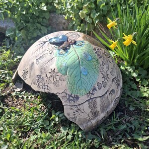 Peut inclure: Un récipient en céramique en forme de dôme avec un motif de feuille. Le récipient est beige avec une feuille verte et des accents bleus. La feuille a un aspect texturé avec des détails de gouttelettes d'eau. Le récipient est entouré d'herbe verte et de feuillage.