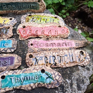 Op de afbeelding: Een set van negen keramische plantenmarkeringen met verschillende namen van kruiden erop geschreven. De markeringen zijn versierd met een bruin en wit patroon en hebben een rustieke uitstraling. De namen van de kruiden zijn: Basilicum, Oregano, Rozemarijn, Wermut, Tijm, Dille, Salie, Lavendel en Bieslook.