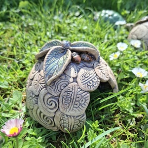 Peut inclure: Sculpture artisanale en céramique représentant un gland stylisé, avec des motifs en relief complexes et des accents de feuilles. La sculpture est dans des tons naturels de terre, avec des détails verts et bruns. L'œuvre est dans un environnement herbeux extérieur.