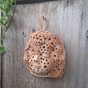 Könnte beinhalten: Ein Terrakotta-Bienenhaus in Form eines Gesichts mit Löchern, in denen Bienen nisten können. Das Gesicht hat geschlossene Augen, eine kleine Nase und einen leicht geöffneten Mund. Das Gesicht ist mit Blumen und Blättern verziert.