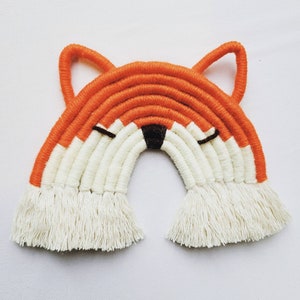 May include: A handmade orange and white yarn fox rainbow wall hanging with a brown nose and eyes. The rainbow is made of thick yarn and has a fringe at the bottom.