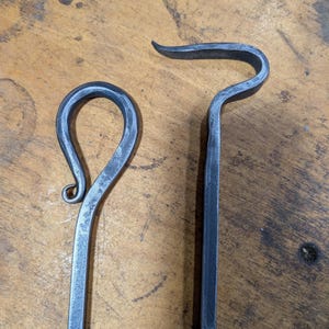 May include: Two hand-forged iron fireplace tools with a dark gray, textured finish. One tool features a circular loop handle with a decorative scroll end, while the other tool has a curved hook top and a twisted metal shaft design.