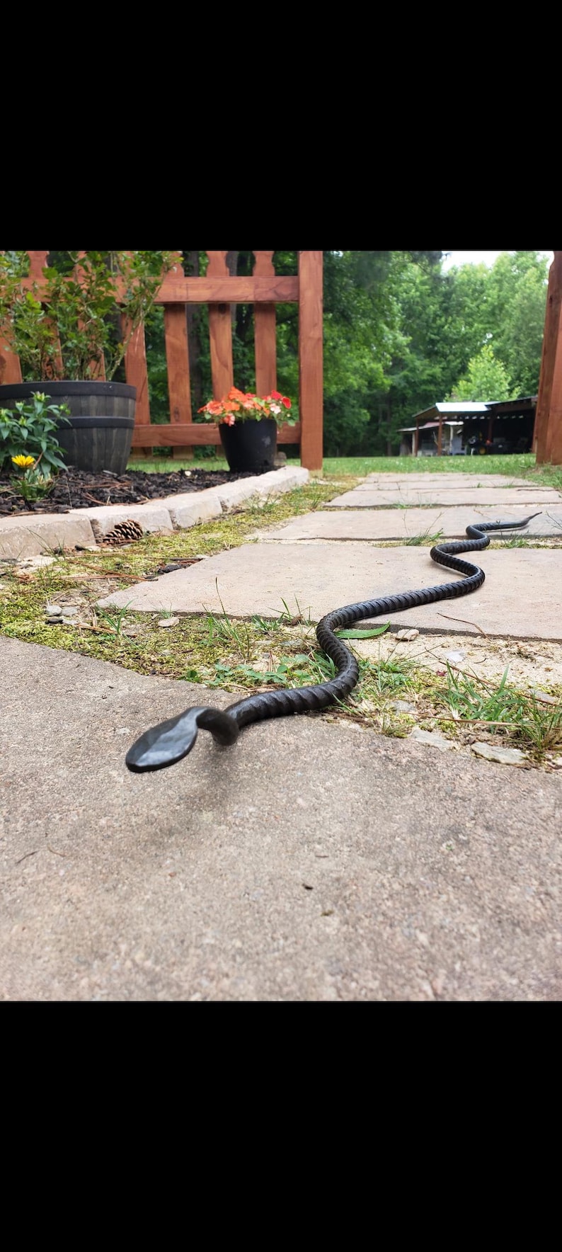 May include: A handcrafted, black metal snake sculpture with a textured body and a flat, spade-shaped head. The snake is positioned on a concrete walkway, with a garden and wooden fence in the background.