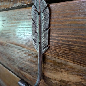 May include: A handcrafted metal wall hook shaped like a feather. The hook is a dark, rustic silver color with detailed feather vein etching. The hook is mounted on a wooden surface with visible wood grain.