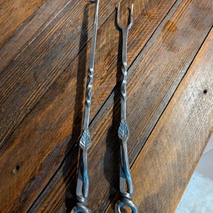 May include: Two hand-forged metal fireplace tools. One is a poker with a pointed end, and the other is a fork-shaped tool. Both have twisted shafts, leaf accents, and looped handles. The tools are made of dark metal.