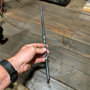 May include: A hand-forged, dark metal wand with a twisted spiral design. The wand tapers to a sharp point at one end and has a rounded base. The metal has a dark, slightly textured appearance, suggesting a blacksmith's craftsmanship.