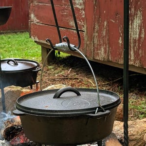 May include: A cast iron Dutch oven with a lid, suspended over a campfire. The pot hangs from a black metal stand with decorative hooks. The fire is burning with visible flames and logs. The background includes a red wooden structure.
