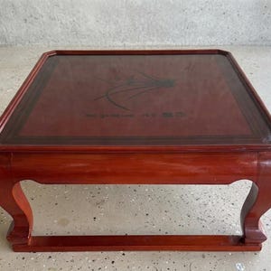 May include: A square low-profile table with a glossy reddish-brown finish. The tabletop features a dark border and a central black ink illustration of a plant with thin leaves, accompanied by black calligraphic text and a small red square seal.