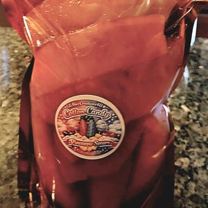 Pode incluir: Um saco de plástico transparente vermelho cheio de pedaços de abacaxi. O saco tem um adesivo redondo com as palavras "Cotton Candy Pineapple Spears" e ilustrações coloridas.