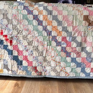 May include: A rectangular patchwork quilt featuring a repeating bow tie pattern composed of numerous small fabric squares in various colors, including red, blue, pink, green, and brown, with a light blue border framing the entire textile piece.