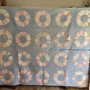 May include: A vintage quilt with a light blue background featuring 15 appliqued ring designs. Each ring is made of various patterned fabric pieces in pastel colors, creating a floral or wreath-like appearance. The quilt is displayed against a wall.