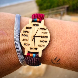 Può includere: Un orologio da polso in legno con un cinturino in pelle marrone e un quadrante bianco. Il quadrante dell'orologio ha la parola "NOW" scritta più volte. L'orologio è al polso di una persona.
