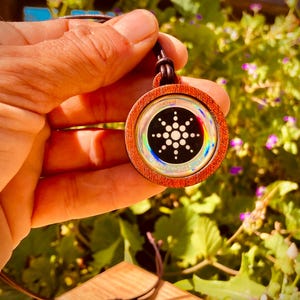 Puede incluir: Un colgante de madera con un diseño circular, iridiscente y holográfico que presenta un círculo central con puntos radiantes. El colgante está en un cordón de cuero marrón y tiene el texto "QUANTUM SCIENCE" alrededor del borde exterior.