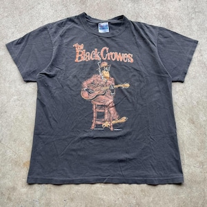 May include: Dark gray t-shirt featuring the text "The Black Crowes" in orange and a cartoon duck playing a guitar. The duck is wearing a suit and hat. The shirt is a short-sleeved crew neck.