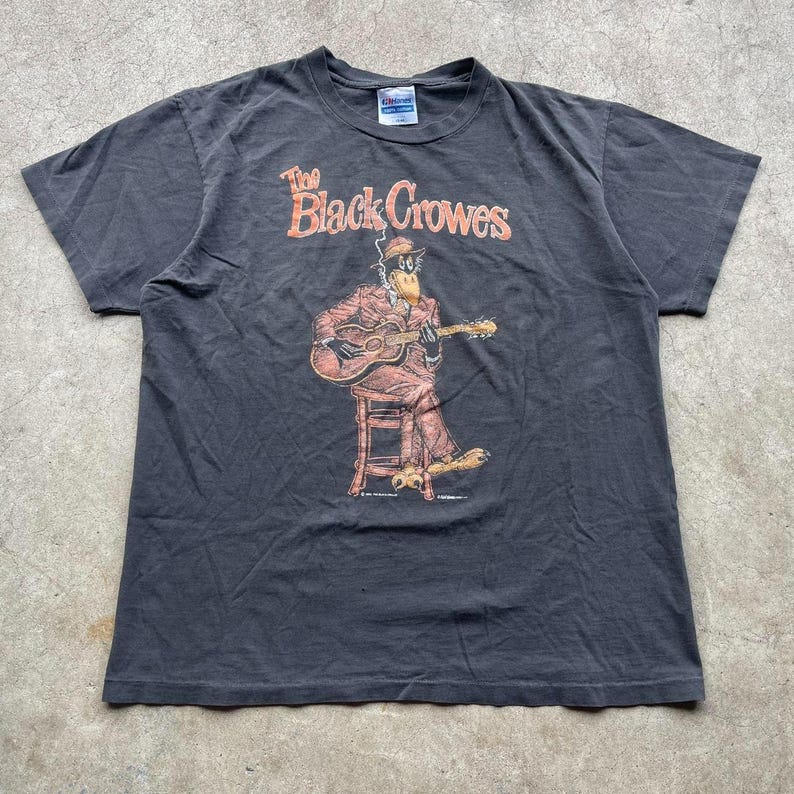 May include: Dark gray t-shirt featuring the text "The Black Crowes" in orange and a cartoon duck playing a guitar. The duck is wearing a suit and hat. The shirt is a short-sleeved crew neck.
