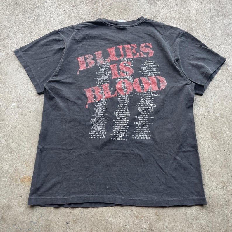 May include: A dark gray t-shirt with the text "BLUES IS BLOOD" in red, along with a list of cities. The shirt has a vintage, worn look, with a crew neck and short sleeves. The text is slightly distressed.
