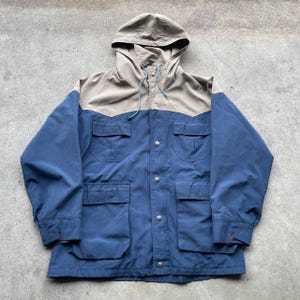 Può includere: Giacca con cappuccio blu e beige con più tasche e chiusure a scatto argentate. La giacca ha un cappuccio con coulisse e un design classico. La giacca è realizzata in materiale resistente.