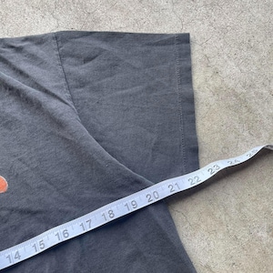 May include: Close-up of a dark gray t-shirt with a measuring tape across the fabric. The tape shows measurements in inches, indicating the size or dimensions of the garment. The shirt's texture and details are visible.