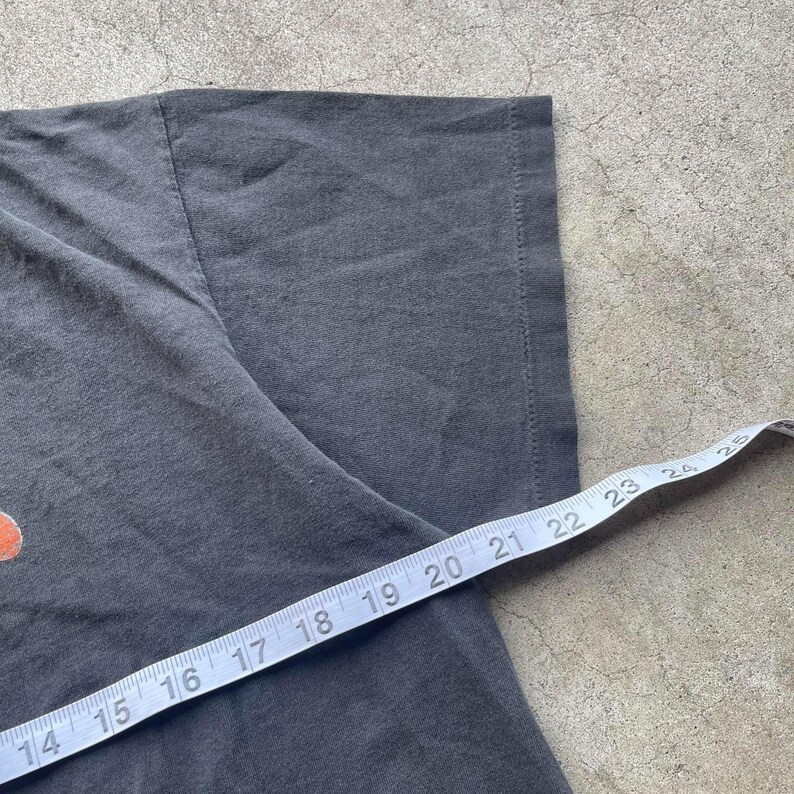 May include: Close-up of a dark gray t-shirt with a measuring tape across the fabric. The tape shows measurements in inches, indicating the size or dimensions of the garment. The shirt's texture and details are visible.