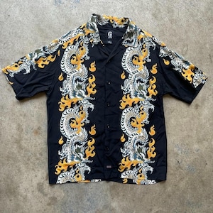 May include: Black button-up shirt with a dragon and flame design. The shirt features a classic collar and short sleeves. The dragon design is white, yellow, and green. The shirt has the brand logo "G" on the collar.