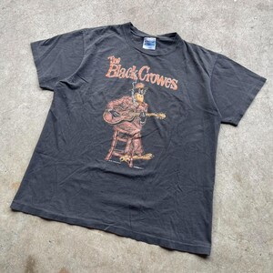 May include: Dark gray t-shirt featuring the text "The Black Crowes" in orange. The graphic depicts a cartoon figure playing a guitar while seated on a stool. The figure is wearing a suit and hat.